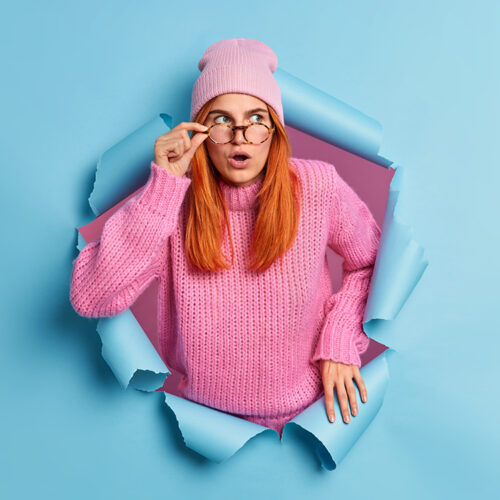 Beautiful surprised redhead young woman keeps mouth opened sees something unbelievable amazes by unexpected offer wears pink hat and sweater breaks through blue paper background. Wow incredible