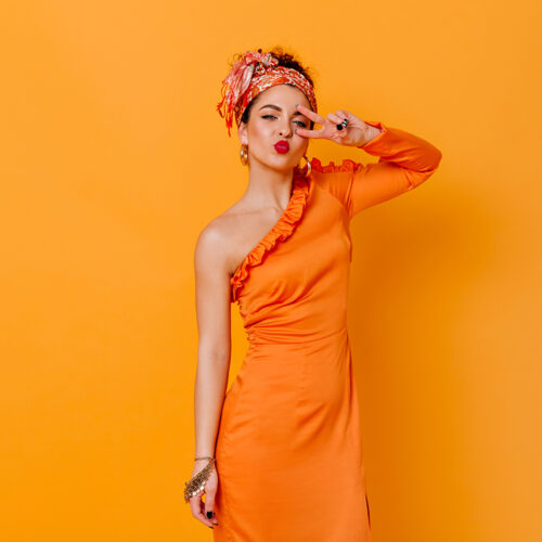 Coquettish woman in silk long dress and headband blows kiss and shows peace sign on orange background
