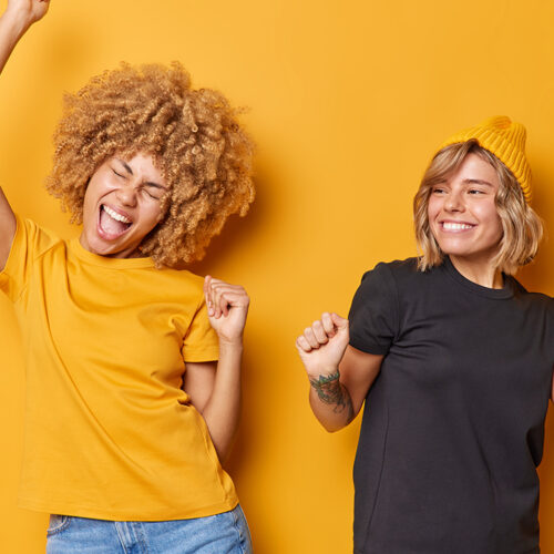 Enjoying life. Carefree positive young friendly women dance and have fun enjoy good time foolish around dressed in casual clothes isolated over yellow background take break move and listen music