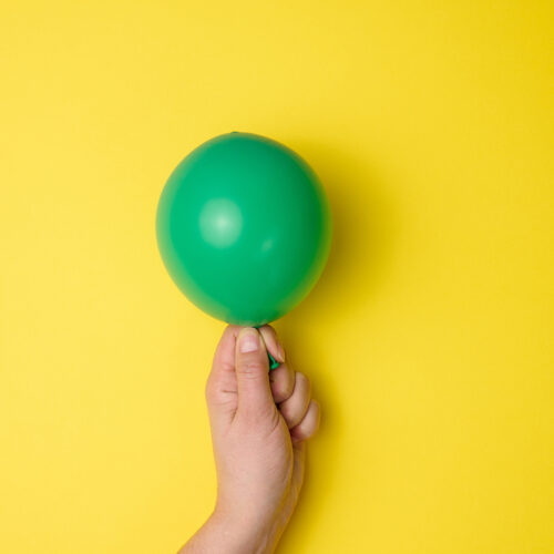 female hand holding an inflated green air balloon on a yellow background, close up