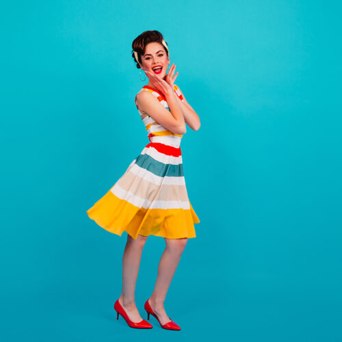 Full length view of pinup girl in striped dress. Studio shot of brunette lady dancing on blue background.