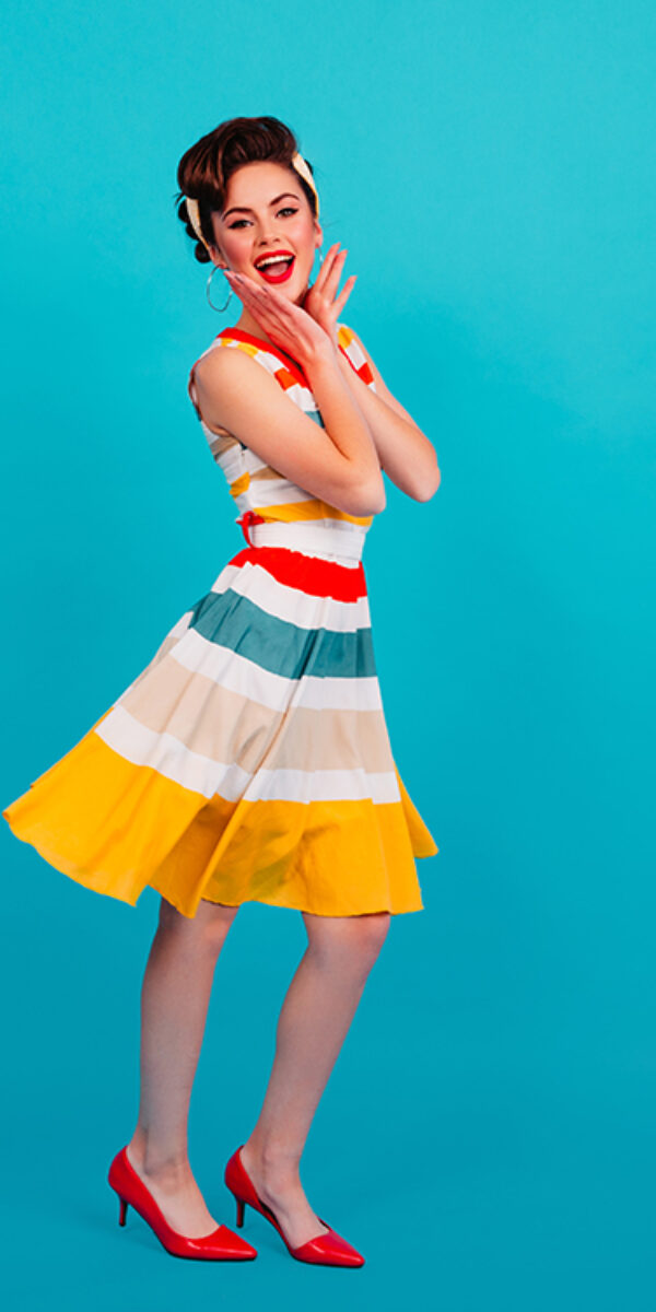 Full length view of pinup girl in striped dress. Studio shot of brunette lady dancing on blue background.