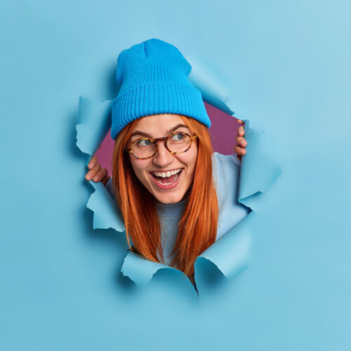 Happy redhead young woman giggles positively and looks aside has fun feels entertained wears optical glasses blue hat looks through torn paper background. Joyful foxy female teenager indoor.