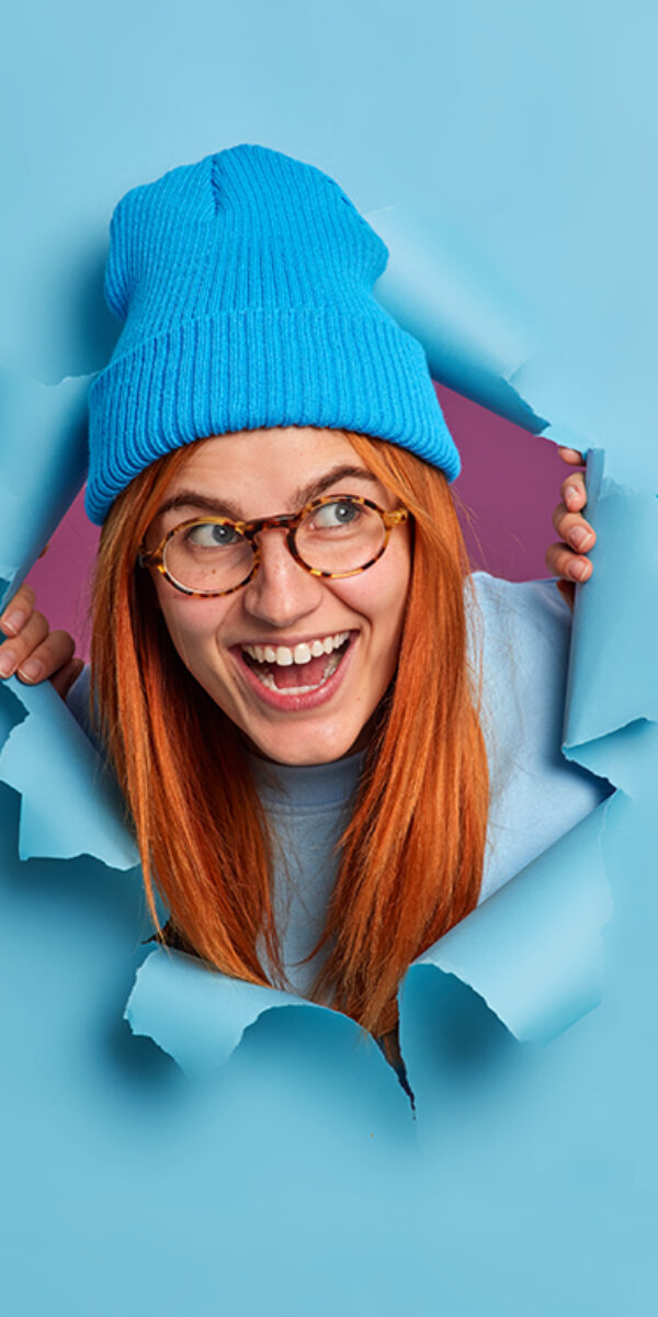 Happy redhead young woman giggles positively and looks aside has fun feels entertained wears optical glasses blue hat looks through torn paper background. Joyful foxy female teenager indoor.