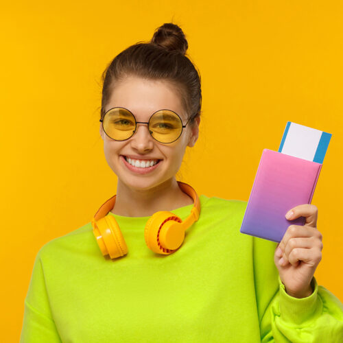 Happy smiling female wearing neon green top, glasses and wireless headphones around neck, holding passport with airplane tickets, isolated on yellow background