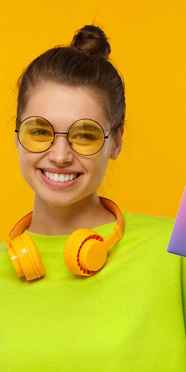 Happy smiling female wearing neon green top, glasses and wireless headphones around neck, holding passport with airplane tickets, isolated on yellow background