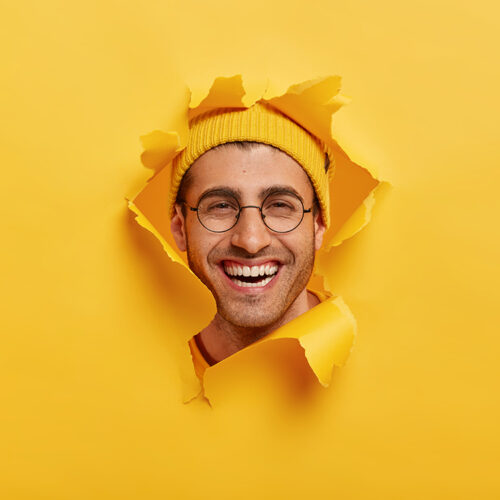 Headshot of positive unshaven young man smiles broadly, wears round optical glasses, yellow headgear, enjoys good time while making photos, looks through torn paper hole, expresses happiness