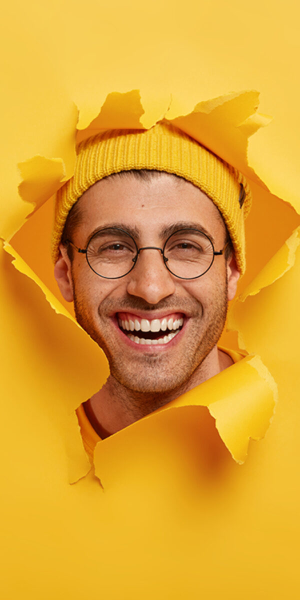 Headshot of positive unshaven young man smiles broadly, wears round optical glasses, yellow headgear, enjoys good time while making photos, looks through torn paper hole, expresses happiness