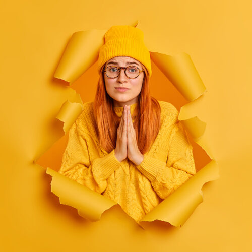 Hopleless sad woman has imploring expression keeps palms together asks for apology wears yellow hat and sweater poses through paper hole of yellow background. Please help me or give chance to improve
