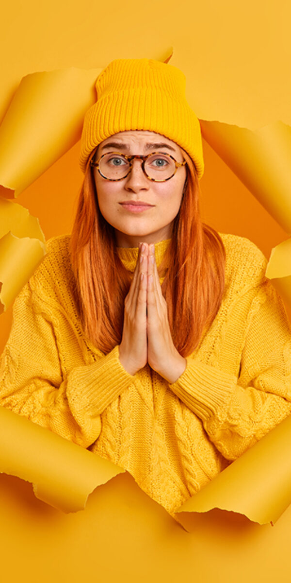 Hopleless sad woman has imploring expression keeps palms together asks for apology wears yellow hat and sweater poses through paper hole of yellow background. Please help me or give chance to improve