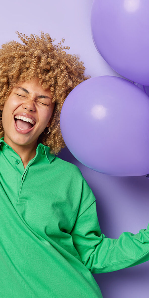Horizontal shot of overjoyed woman keeps palm raised up holds bunch on inflated balloons dressed in green jumper isolated over purple background celebrates anniversary. Happy birthday concept