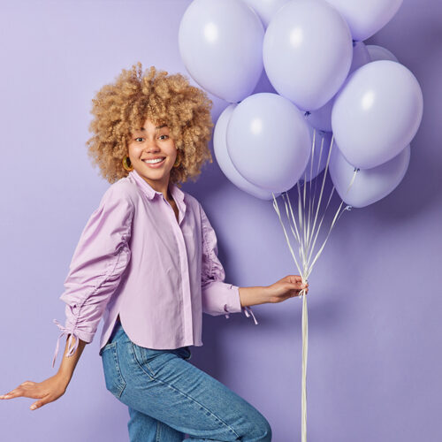 Horizontal shot of positive woman with curly bushy hair holds bunch of inflated balloons celebrates special occasion dressed in shirt and jeans poses against purple background. Holiday concept