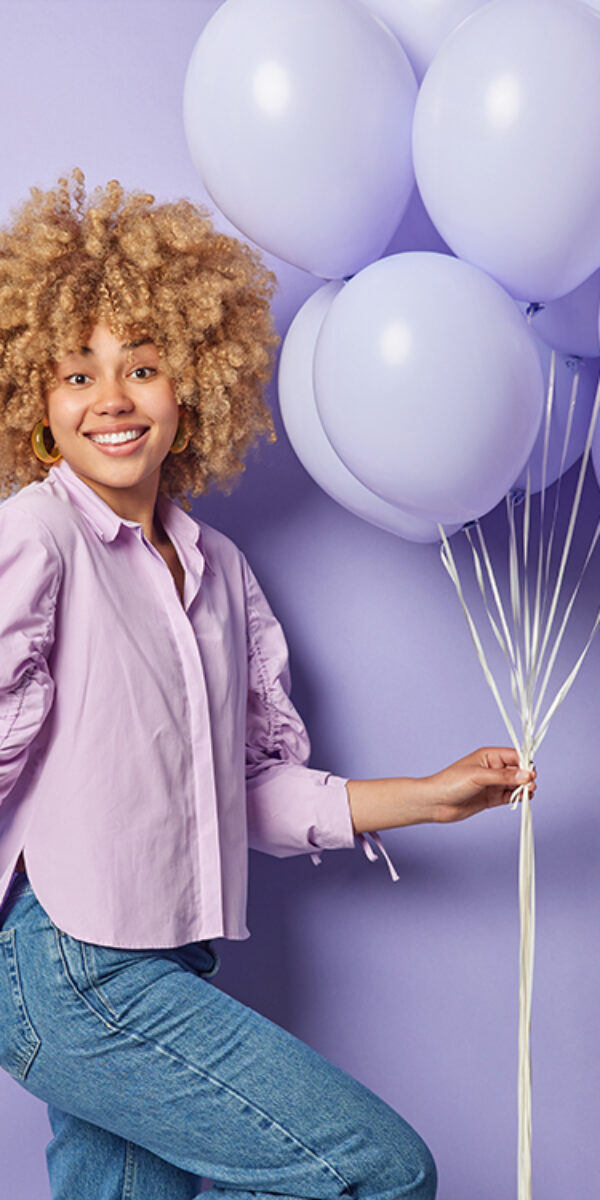 Horizontal shot of positive woman with curly bushy hair holds bunch of inflated balloons celebrates special occasion dressed in shirt and jeans poses against purple background. Holiday concept