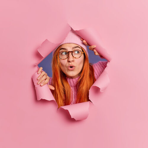 Impressed redhead European woman keeps mouth opened from amazement reacts on seeing something shocking wears hat and spectacles breaks through paper pink background. Omg and reaction concept
