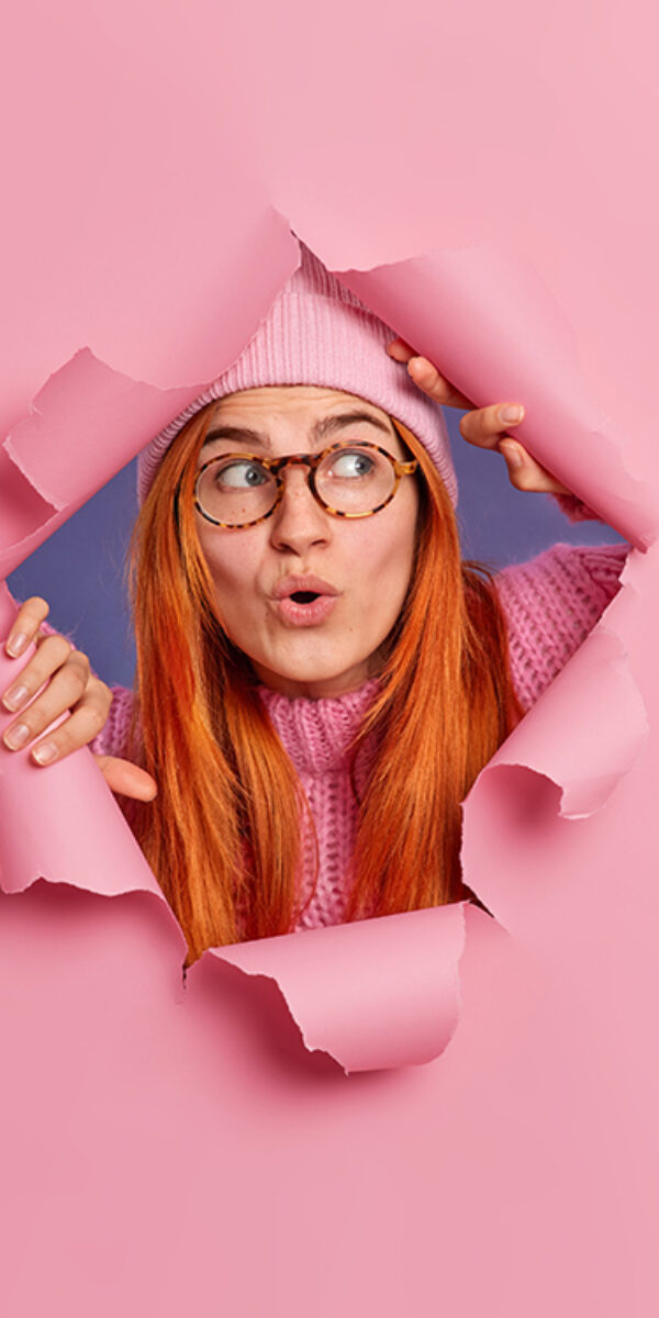 Impressed redhead European woman keeps mouth opened from amazement reacts on seeing something shocking wears hat and spectacles breaks through paper pink background. Omg and reaction concept