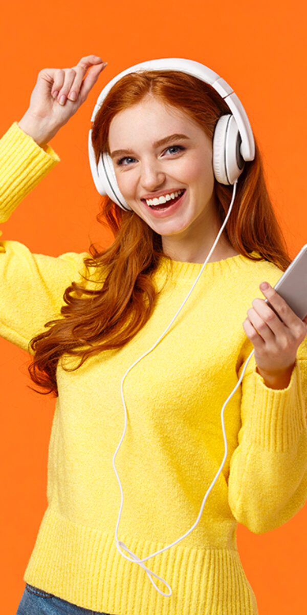 Lets dance. Cheerful carefree modern hipster girl with red curly hair and freckles, dancing lift hand up and holding smartphone, listen music with new ordered online headphones, orange background.
