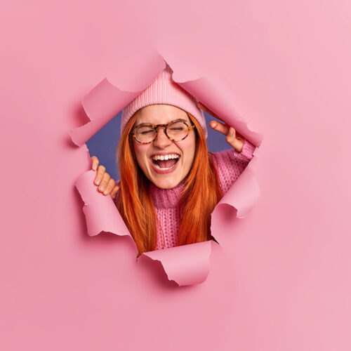 Overjoyed redhead freckled woman wears glasses and hat poses through ripped hole laughs joyfully keeps mouth opened keeps eyes closed. Satisfied foxy European girl hears something very funny