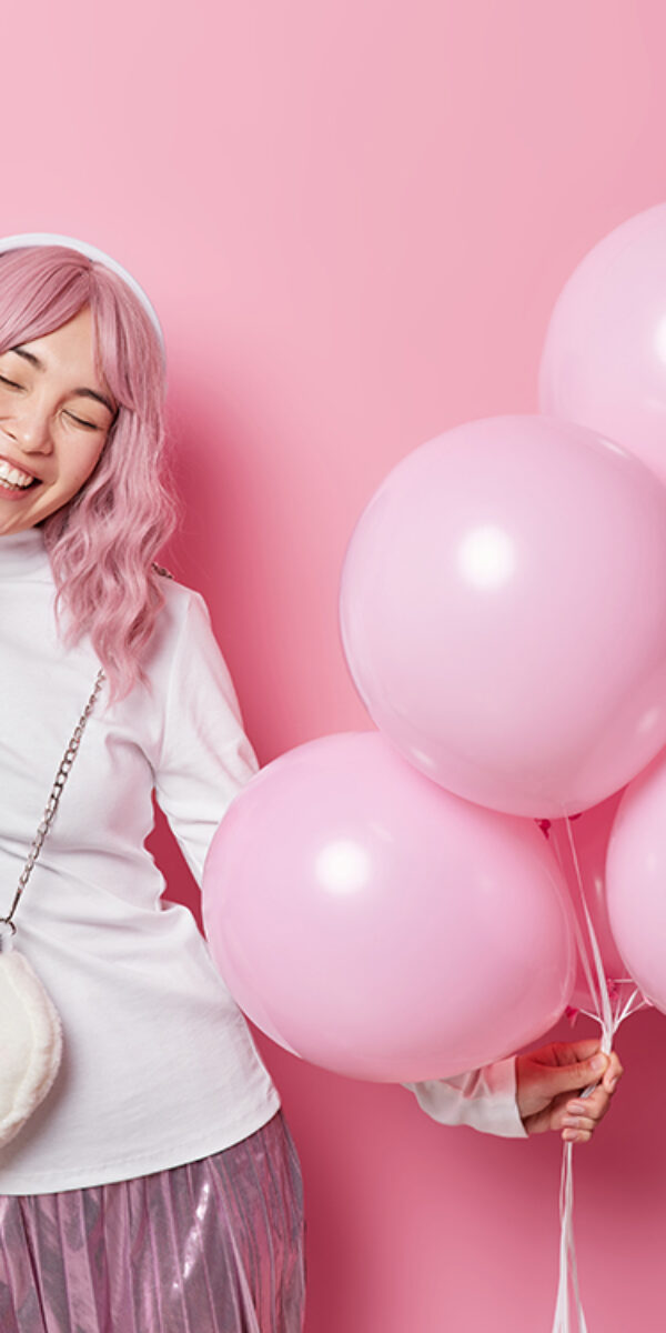 Positive long haired millennial girl listens favorite music holds bunch of inflated balloons carries bag wears white turtleneck and skirt has festive mood isolated on pink background. Holiday concept