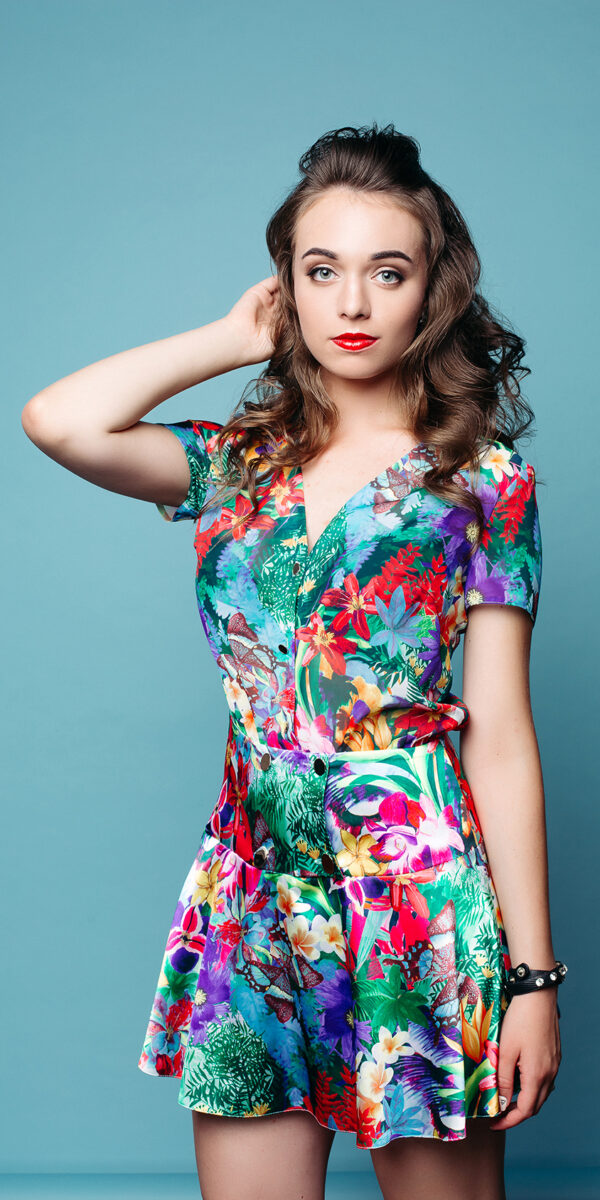 Studio portrait of beautiful young girl with red lips and wavy hair wearing lovely floral dress with hand at hair looking at camera over azure background.