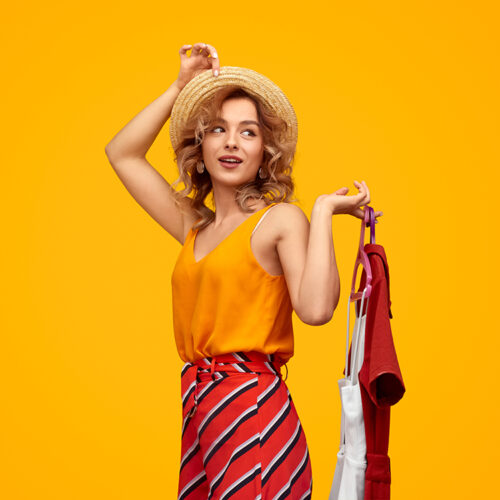 Happy young woman in straw hat looking away and holding hangers with stylish garments against vivid yellow background