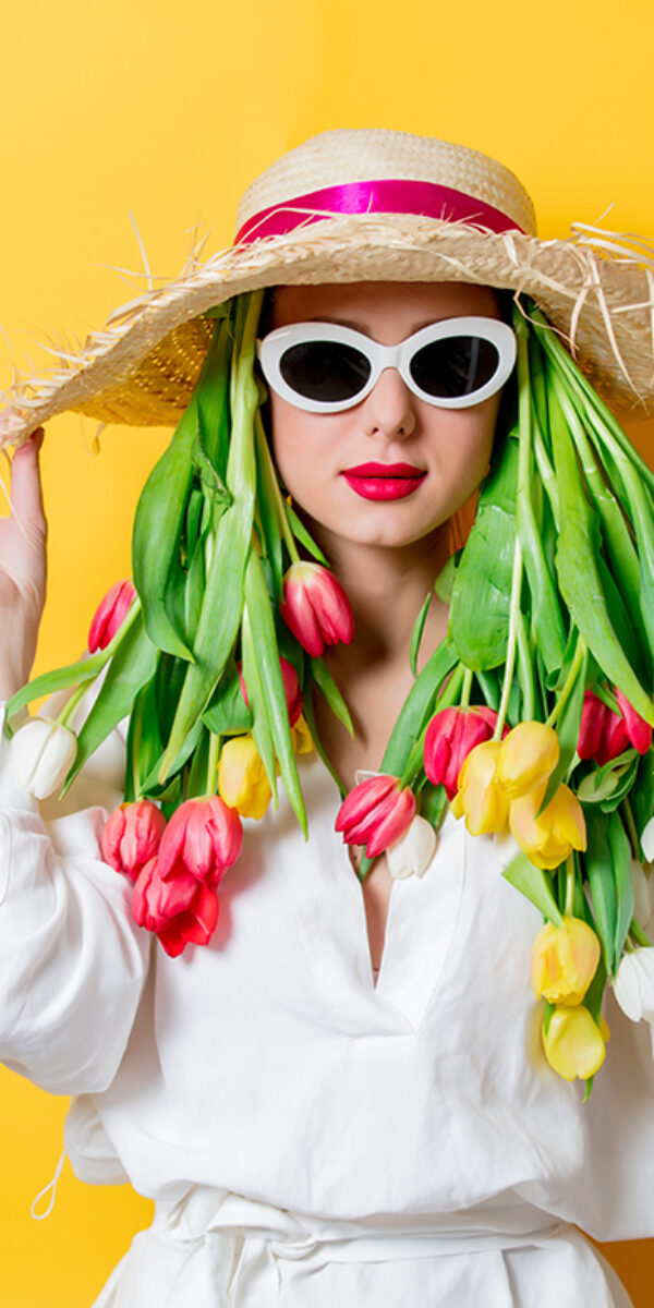 Beautiful woman in white shirt and hat with fresh springtime tulips and sunglasses on yellow background
