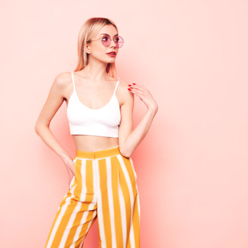 Young beautiful smiling blond female in trendy summer white top t-shirt and yellow stripped pants clothes. Sexy carefree woman isolated on pink in studio. Positive model having fun. In sunglasses