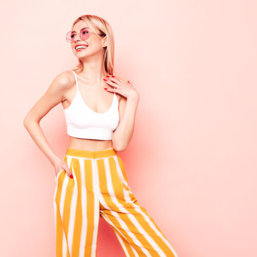 Young beautiful smiling blond female in trendy summer white top t-shirt and yellow stripped pants clothes. Sexy carefree woman isolated on pink in studio. Positive model having fun. In sunglasses