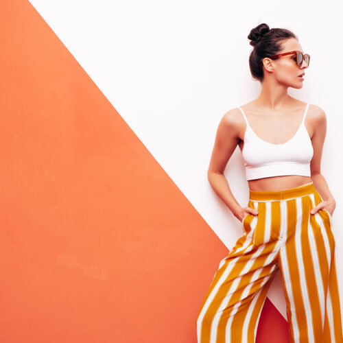 Fashion young beautiful smiling female in trendy summer white top t-shirt and yellow striped pants. Sexy carefree woman posing near wall in studio interior. Positive model. In sunglasses
